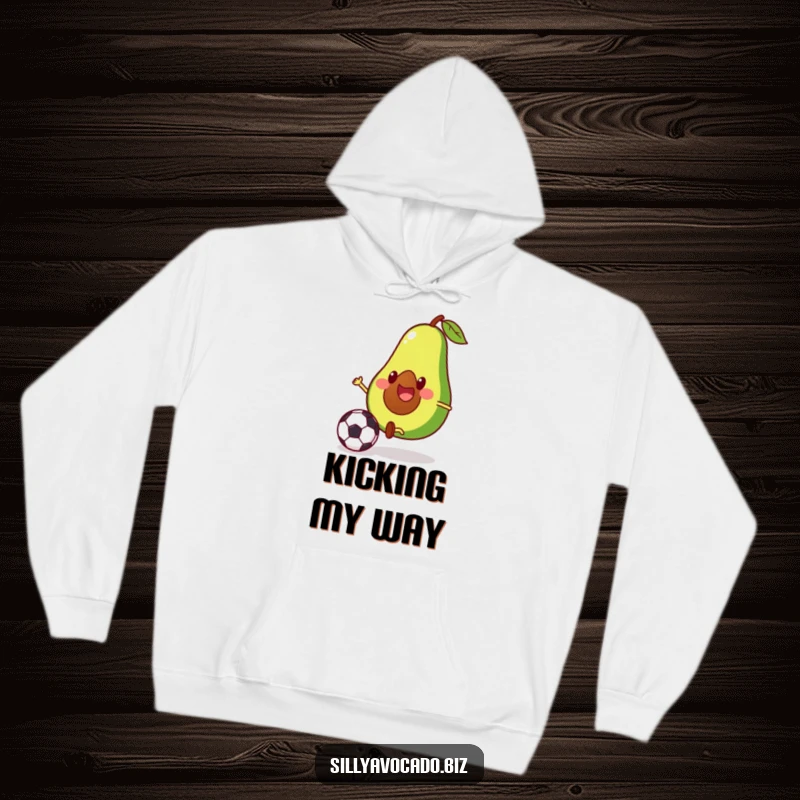 Funny hoodie featuring a smiling avocado with tiny legs focused on kicking a soccer ball, perfect for sports lovers.